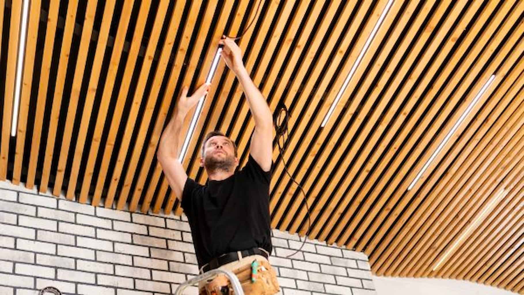 A male electrician installing strip lighting on a wood slat ceiling.