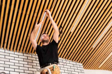 A male electrician installing strip lighting on a wood slat ceiling.